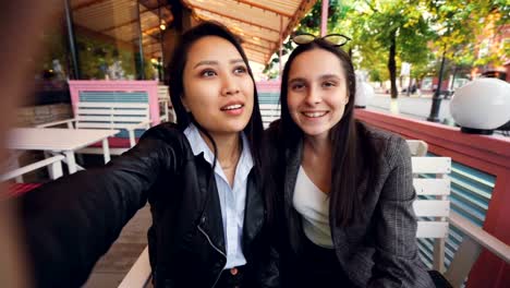 Point-of-view-shot-of-happy-female-friends-making-online-video-call-and-talking-sitting-in-street-cafe-together-showing-milkshake-glasses-and-having-fun.