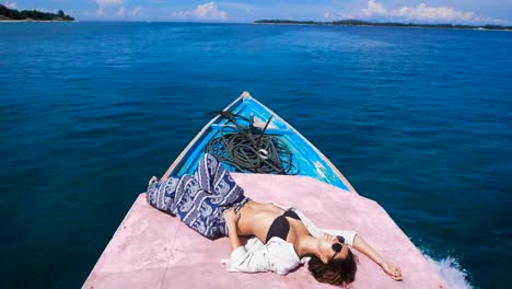 Tanned-young-girl-in-sunglasses-with-a-thrown-back-arm-behind-her-head-is-in-the-stern-of-the-boat,-which-quickly-goes-on-the-dark-blue-ocean-to-a-small-island