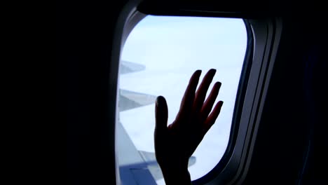 Vista-desde-la-ventana-del-avión,-se-puede-ver-la-oscura-silueta-de-la-mano-femenina,-la-palma,-el-movimiento-de-los-dedos,-el-cielo,-las-nubes,-el-ala-del-avión.-El-tiempo-es-claro,-soleado