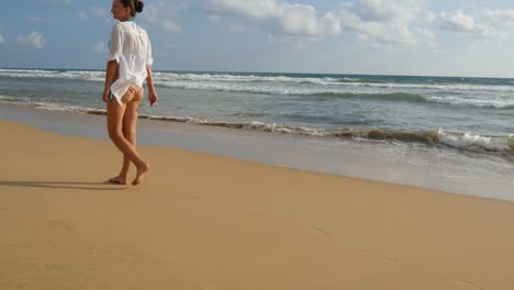 Beautiful-woman-in-swimsuit-and-shirt-walking-on-sea-beach-barefoot.-Young-girl-going-on-the-ocean-shore.-Female-foot-stepping-on-the-sand-with-sea-waves-background.-Summer-vacation-concept-Rear-view