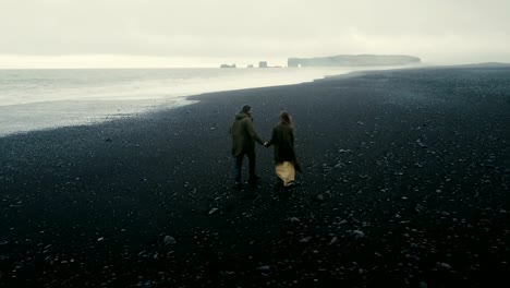 Back-aerial-view-of-the-young-couple-walking-on-the-black-volcanic-beach-in-Iceland.-Romantic-date-near-the-sea