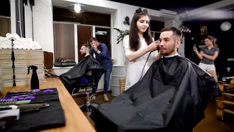 Zwei-Freunde-besuchen-moderne-Barbershop.-Hübsche-Hipster-gegen-Spiegel-sitzen-und-arbeiten-mit-Friseuren.-Lächelnder-Mann-und-Frau-mit-professioneller-Ausrüstung-für-die-Herstellung-von-stilvollen-Haarschnitte