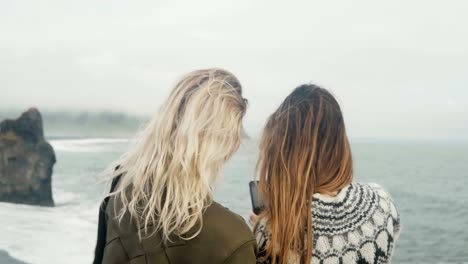 Two-happy-women-taking-selfie-photos-on-smartphone,-girls-spending-time-on-the-beach-near-the-troll-toes-in-Iceland