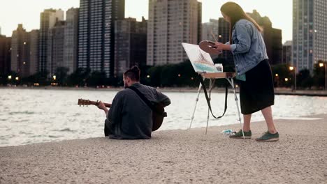 Joven-pareja-creativa-de-artistas-callejeros-haciendo-afición-en-orilla-de-Michigan,-Chicago,-Estados-Unidos.-Sorteos-de-mujer,-hombre-juegos-de-guitarra