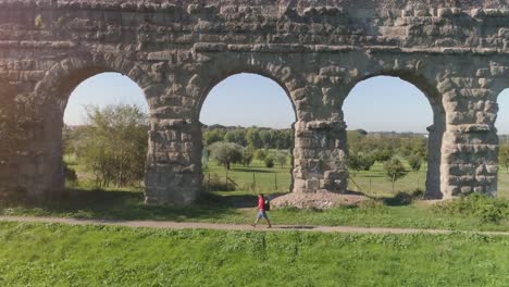 Hombre-joven-mochilero-caminando-por-camino-de-tierra-a-lo-largo-del-antiguo-acueducto-romano-en-naranja-ropa-deportiva-senderismo-dolly-de-drone-de-vista-aérea