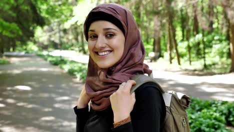 Portrait-of-a-young-smiling-girl-in-a-hijab-standing-in-park-with-a-backpack-traveling-concept-50-fps
