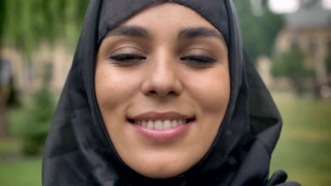 Cara-de-joven-hermosa-chica-musulmana-en-hijab-es-de-pie-y-sonriente-durante-el-día,-mal-tiempo,-mirando-a-cámara,-concepto-de-epoca,-fondo-borroso