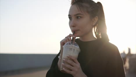 Young-brunette-is-drinking-ice-latte-through-straw-in-quay-in-evening