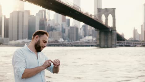 Erfolgreicher-junger-Geschäftsmann-Stand-in-der-Nähe-Fluss-unter-Brooklyn-Bridge,-mit-seiner-smart-Uhr,-zu-Fuß-entfernt-4K