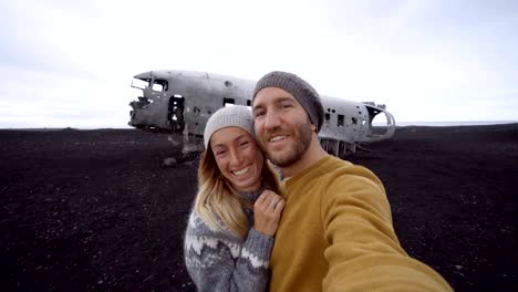 Junges-paar-stehend-durch-Flugzeug-Wrack-am-schwarzen-Sandstrand-unter-Selfie-Porträt-Sehenswürdigkeit-zu-besuchen-in-Island-und-stellen-mit-dem-Wrack---4K