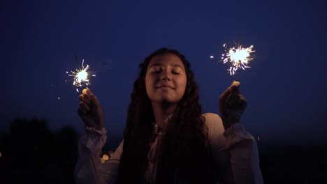 Ein-junges-Mädchen-mit-langen,-dunklen-Haaren-steht-vor-dem-Hintergrund-der-Stadt-in-der-Nacht-und-Feuerwerk-bei-Laune-hält.-Slow-Motion.-4K