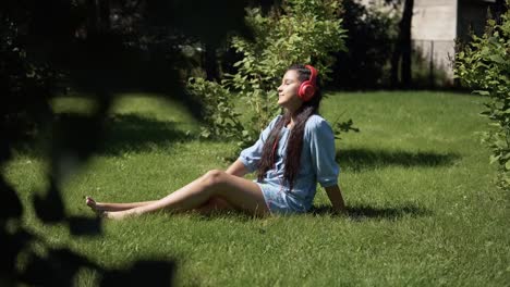 Young-girl-listening-to-music-on-headphones-sitting-on-grass-in-park-in-sunny-weather.-4K