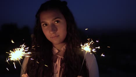 Ein-junges-Mädchen-mit-langen,-dunklen-Haaren-steht-vor-dem-Hintergrund-der-Stadt-in-der-Nacht-und-Feuerwerk-in-ihren-Händen-hält.-Slow-Motion.-4K