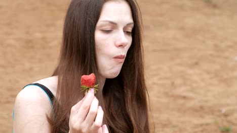 Young-woman-brunette-eats-a-strawberry-sitting-on-the-beach.-Face-close-up.