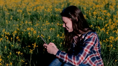 Woman-looking-at-mobile-phone-sitting-in-Park-on-grass-among-yellow-flowers.