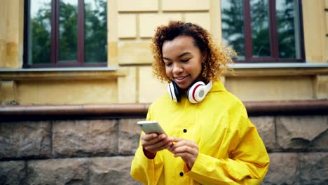 Happy-African-American-student-is-using-smartphone-browsing-or-texting-friends-standing-outdoors-in-the-street-of-modern-city-wearing-headphones-and-smiling.