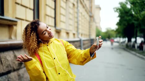 Niedlicher-afrikanischen-amerikanischer-Teenager-spricht-mit-Freunden-online-Skypen-mit-Smartphone-und-am-Bildschirm-stehen-draußen-in-der-Straße-und-tragen-Kopfhörer-suchen.