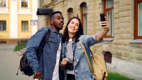 Happy-tourists-friends-with-backpacks-are-taking-selfie-using-smartphone-showing-hand-gestures-v-sign-and-thumbs-up-standing-together-in-the-street-in-beautiful-city.