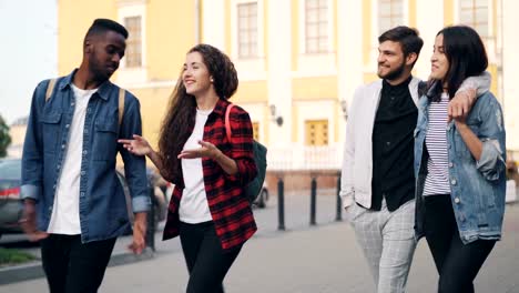 Friends-tourists-in-casual-clothes-are-walking-in-city-with-backpacks-talking-and-laughing-on-summer-day.-Friendship,-young-people-and-tourism-concept.