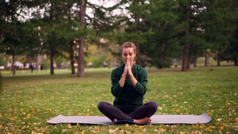 Hübsche-junge-Frau-ist-finishing-Yoga-Praxis-auf-Matte-Augen-schließen-und-atmen-an-friedlichen-Herbsttag-im-Park-sitzen.-Freizeitbeschäftigung-und-urbane-Menschen-Konzept.