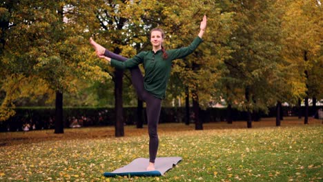 Smiling-girl-professional-yoga-instructor-is-doing-balancing-exercises-standing-on-one-leg-on-mat-on-grass-in-park.-Beautiful-autumn-nature-is-visible.