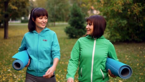 Dolly-shot-of-good-looking-sportswomen-talking,-smiling-and-holding-yoga-mats-walking-in-park-on-autumn-day.-Urban-people,-leisure-and-sports-concept.