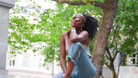 Moda-joven-mujer-de-negra-en-gafas-de-sol,-camiseta-y-lateral-banda-vaqueros-sentados-cerca-de-árbol-en-la-calle-de-la-ciudad