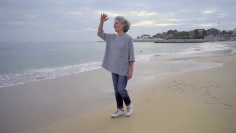 Glückliche-ältere-Frau-tragen-Freizeitkleidung-und-Turnschuhe-zu-Fuß-auf-nassen-Sand-und-Seawaves.