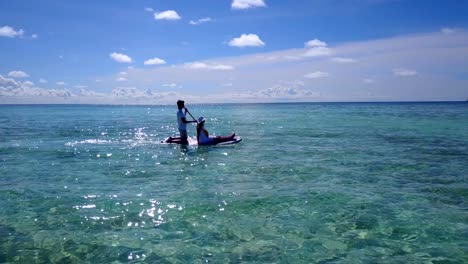 v03860-fliegenden-Drohne-Luftaufnahme-der-Malediven-weißen-Sandstrand-2-Personen-junges-Paar-Mann-Frau-entspannend-auf-Paddleboard-auf-sonnigen-tropischen-Inselparadies-mit-Aqua-blau-Himmel-Meer-Wasser-Ozean-4k