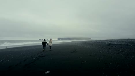 Aerial-back-view-of-the-young-couple-in-icelandic-sweater-walking-on-shore-of-the-sea-on-black-volcanic-beach-in-Iceland