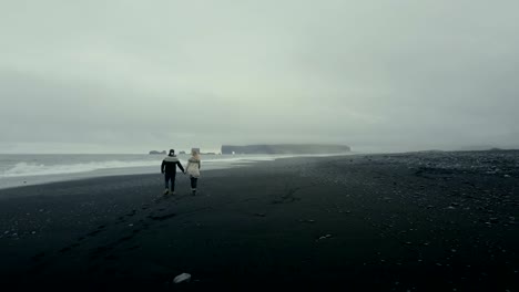 Vista-aérea-de-espalda-de-la-joven-pareja-en-Jersey-islandés-caminando-por-la-orilla-del-mar-en-la-playa-volcánica-negra-en-Islandia