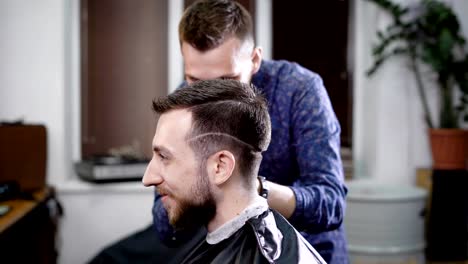 Side-view-of-a-head-of-stylish-smiling-bearded-man-sitting-in-the-barbershop-covered-with-black-peignoir.-Barber-in-casual-clothes-standing-behind-customer-making-boxer-haircut-with-clipper-and-comb