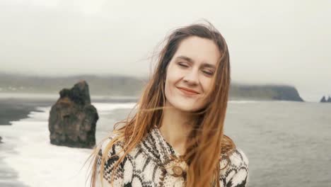 Portrait-of-young-happy-woman-standing-on-black-beach,-looking-on-camera-and-smiling,-near-troll-toes-rocks-in-Iceland