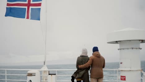 Back-view-of-young-stylish-couple-standing-on-the-board-of-the-ship-with-Icelandic-flag.-Man-and-woman-look-on-the-sea