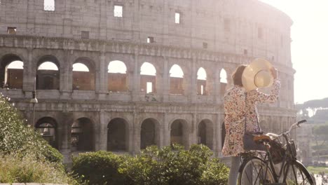 Hermosa-mujer-joven-en-moda-colorido-vestido-solo-en-colina-con-bicicleta-mirando-en-el-Coliseo-de-Roma-al-atardecer-con-chica-atractiva-de-árboles-con-elegante-sombrero-de-paja