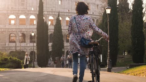 Schöne-junge-Frau-in-bunte-Mode-Kleid-mit-Fahrrad,-zu-Fuß-in-Richtung-Kolosseum-in-Rom-bei-Sonnenuntergang-mit-Bäumen-glücklich-touristisch-attraktiven-Mädchen-in-Colle-oppio