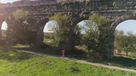 Hombre-joven-mochilero-caminando-por-camino-de-tierra-a-lo-largo-del-antiguo-acueducto-romano-en-naranja-ropa-deportiva-senderismo-dolly-de-drone-de-vista-aérea