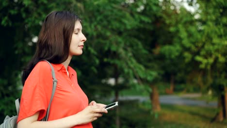 Side-view-of-attractive-pregnant-woman-walking-in-park-and-using-smart-phone,-person-is-watching-screen-then-looking-around-enjoying-beautiful-nature.