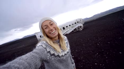 4K-joven-por-ruina-del-aeroplano-en-la-playa-de-arena-negra,-produciendo-una-selfie-retrato-famoso-para-visitar-en-Islandia-y-posan-junto-a-los-restos-del-naufragio