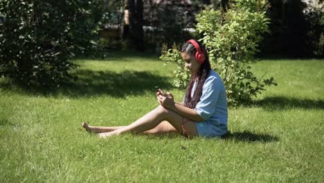 Young-girl-listening-to-music-with-headphones-sitting-on-the-grass-in-the-park.-4K