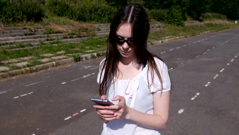 Young-brunette-girl-walking-in-the-stadium-and-typing-a-message-on-a-mobile-phone-and-smiling.