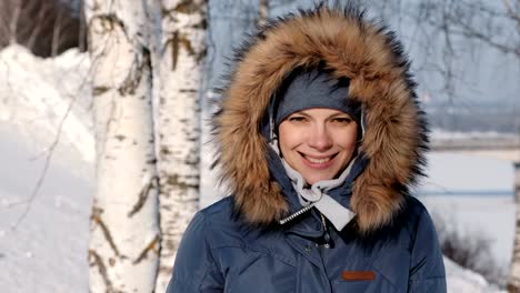 Close-up-Frau-in-blau-Daunenjacke-mit-Fell-Kapuze-Stirnrunzeln-vor-der-Sonne-und-lächelt,-in-Winter-Park