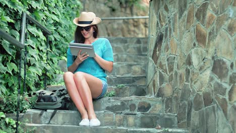 Tiro-completo-bonita-a-mujer-turista-relajada-sentado-en-las-escaleras-con-escalones-de-piedra-con-tablet-PC