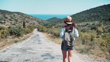 Mujer-de-backpacker-activo-de-vista-posterior-con-sombrero-caminando-en-camino-al-mar-rodeado-de-montañas