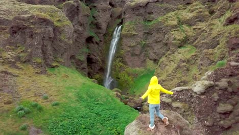 Drone-aerial-view-of-Young-woman-arms-outstretched-at-spectacular-waterfall-in-Iceland--4K-video