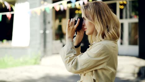 Cheerful-young-girl-is-adjusting-camera-then-taking-photos-of-beautiful-city-standing-outdoors-in-the-street.-Hobby,-occupation,-modern-technology-concept.