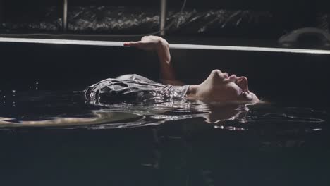 Young-woman-lying-on-smooth-surface-water-in-swimming-pool-on-dark-background