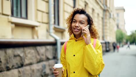 Cheerful-mixed-race-teenage-girl-is-talking-on-smart-phone-and-holding-take-out-coffee-walking-along-street-in-beautiful-city.-Modern-lifestyle-and-technology-concept.