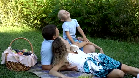 Family-picnic-in-nature.-Mom,-dad-and-son-lie-on-the-grass-in-the-Park-near-the-fruit-basket.-They-look-up,-laugh,-play-and-have-fun.-Boy-eating-grapes.