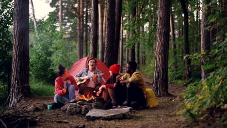 Man-tourist-in-casual-clothing-is-playing-the-guitar-while-his-friends-are-cooking-and-eating-sweet-marshmallow-sitting-around-campfire-on-autumn-day.-People-and-camp-concept.